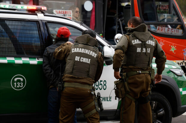 Dos funcionarios de Carabineros de Chile de espaldas proceden a la detención de una persona con gorra roja junto a una patrulla policial.