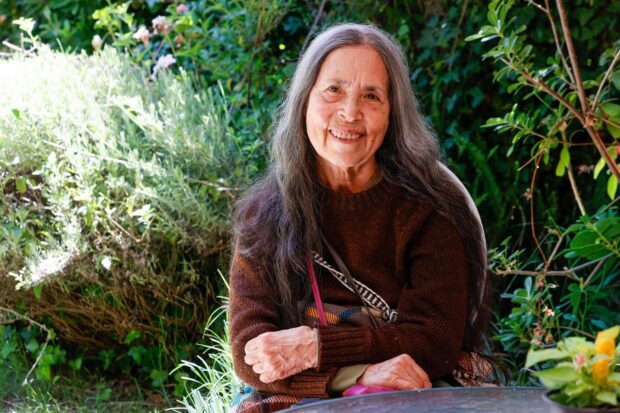 Retrato de Cecilia Vicuña, poeta y artista visual chilena, sentada en un entorno natural, sonriendo, autora de QUIPOema y Precario y reconocida internacionalmente por su obra artística y poética.