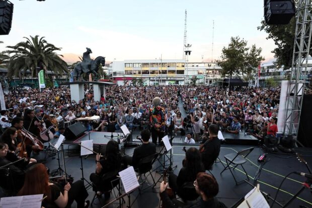 Vista desde el escenario de una orquesta sinfónica tocando ante una gran multitud en una plaza pública, con una estatua ecuestre y edificios de fondo.