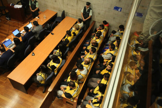 Fotografía tomada desde arriba de más de 25 personas sentadas en tres banquillos de un jurado, todos vestidos con chaleco amarillo.