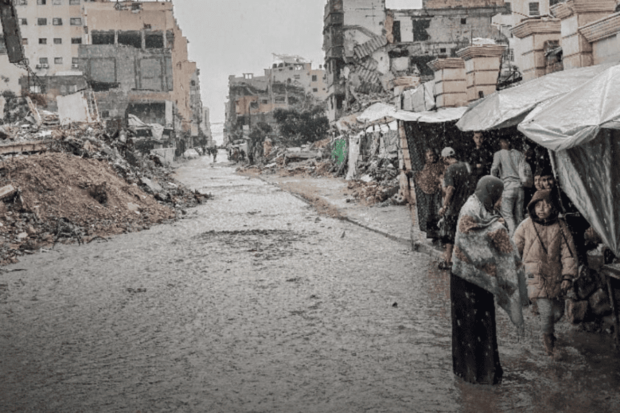 Una calle inundada de barro y escombros en Gaza, flanqueada por edificios destruidos y refugios improvisados, con personas caminando bajo la lluvia.