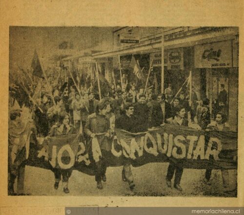 Fotografía histórica en blanco y negro que muestra una manifestación masiva del MIR en una calle urbana. En primer plano, jóvenes sostienen un lienzo oscuro con la frase "AHORA A CONQUISTAR". La multitud camina entre locales comerciales portando banderas. En la base de la imagen se observa el sello de agua de memoriachilena.cl.