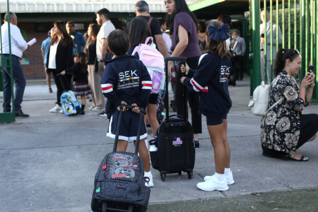 Niños y niñas con mochilas y maletas escolares entrando a un colegio junto a apoderados.