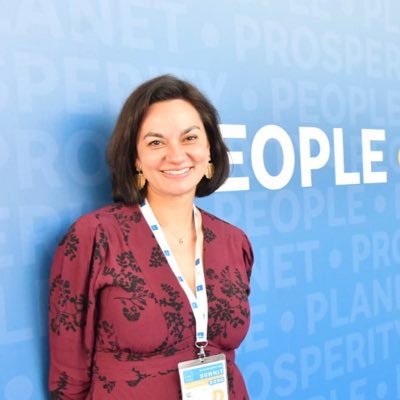 Mujer sonriente frente a un fondo azul con palabras relacionadas a desarrollo y sostenibilidad, usando credencial de conferencia internacional.