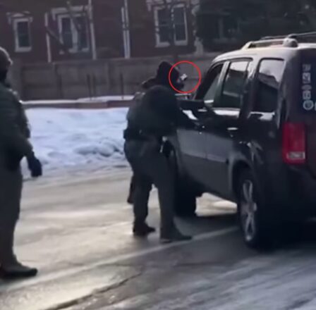 Agente de ICE frente a un vehículo oscuro en una calle con nieve, apuntando con un arma hacia el interior de la ventana del conductor.