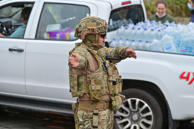 Militar se ve dirigiendo el tráfico. Detrás de él pasa una camioneta pick up blanca llena de botellas de agua.