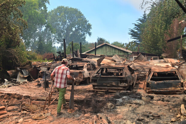Un hombre camina entre varios esqueletos de automóviles calcinados, estructuras de viviendas destruidas y escombros de una propiedad devastada por un incendio forestal.