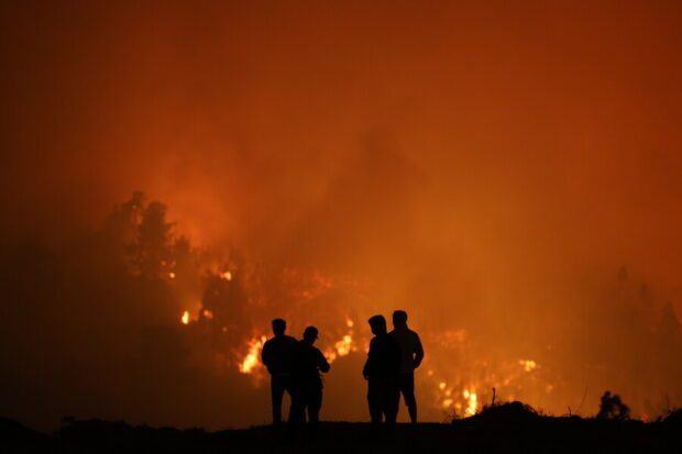 Siluetas de cuatro personas observando a la distancia un incendio forestal de gran magnitud durante la noche.