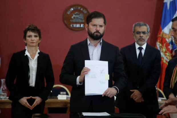 Presidente Boric, junto a la entonces ministra Toha y el ministro Cordero en un cuarto de paredes rojas. Boric sostiene un papel mientras ve con cara seria a la cámara.