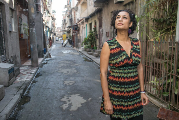 Marielle Franco de pie en una calle estrecha de un barrio popular de Río de Janeiro, mirando hacia arriba con gesto reflexivo.