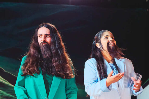Dos intérpretes en escena con trajes de colores y barbas postizas, uno con chaqueta verde y otro celeste, bajo iluminación teatral, en un momento expresivo del montaje.