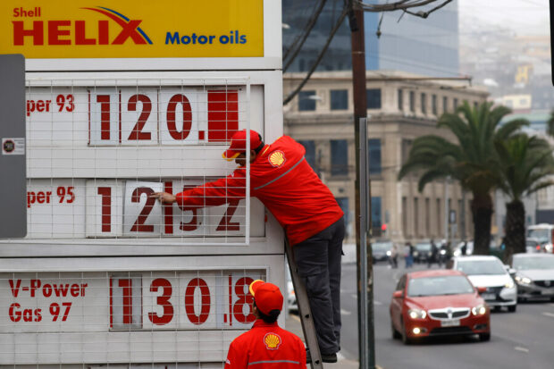 Trabajador de la Shell vestido de rojo mientras cambio los precios del cartel de una bencinera. El cartel es blanco con los números en rojo.