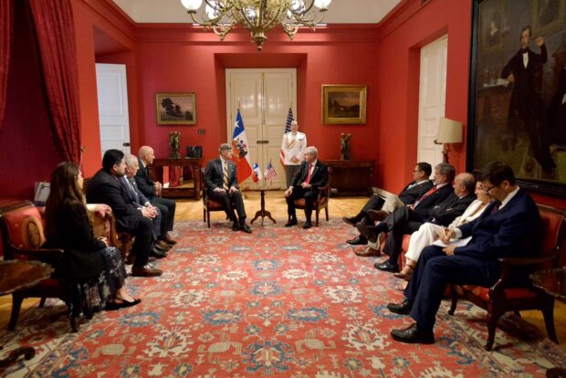 En un salón rojo y con alfombras estambadas, personas vestidas de traje formal, hombres en su mayoría, se sientan en forma de U viendo a la cámara. Al fondo se ven las banderas de Chile y Estados Unidos.