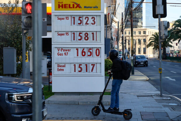 Precios de bencina en estación de servicio Shell con persona en scooter eléctrico pasando por delante.