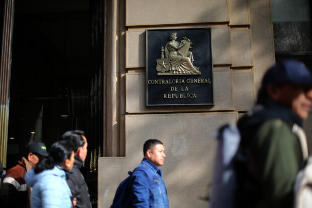 Entrada de la Contraloría General de la República. Se ve un edificio con el escudo del organismo al lado de su puerta. En la foto se ven transeuntes caminando delante del edificio.