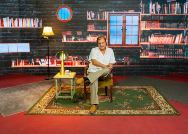 Francisco Vidal sentada en una silla en la mitad de un set de grabación que simulta un departamento con una ventana y repisas con libros.