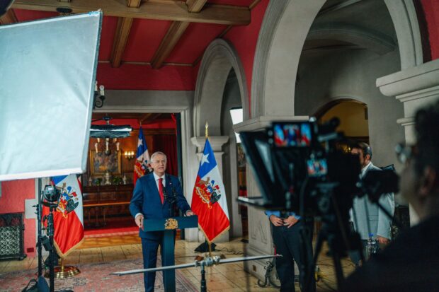 José Antonio Kast frente a un podio con el escudo nacional durante la grabación de una cadena nacional. La toma incluye cámaras de televisión y equipos de iluminación en un salón oficial con banderas chilenas de fondo.