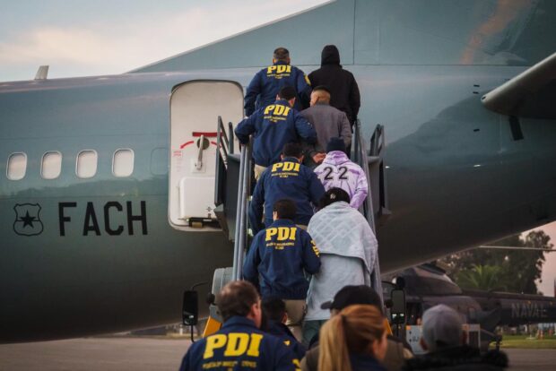 Oficiales PDI custodian fila de personas expulsadas del país mientras suben un avión de la Fuerza Aérea de Chile.