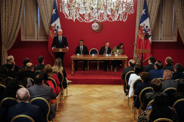 José Antonio Kast hablando en un podio con el escudo nacional de Chile, durante una sesión oficial en un salón del Palacio de La Moneda. Al fondo se observa una bandera chilena y una mesa con autoridades frente a una audiencia.