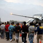 Oficiales PDI custodian fila de personas expulsadas del país mientras se dirigen a un avión de la Fuerza Aérea de Chile en la losa de un aeropuerto.
