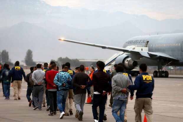Oficiales PDI custodian fila de personas expulsadas del país mientras se dirigen a un avión de la Fuerza Aérea de Chile en la losa de un aeropuerto.