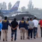 Comisión mixta llegó a acuerdo por proyecto sobre expulsiones de migrantes Oficiales PDI custodian fila de personas expulsadas del país mientras se dirigen a un avión de la Fuerza Aérea de Chile en la losa de un aeropuerto.