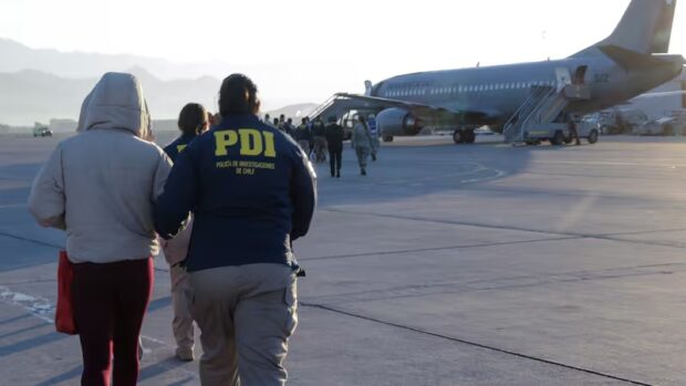 Oficiales PDI custodian fila de personas expulsadas del país mientras se dirigen a un avión de la Fuerza Aérea de Chile en la losa de un aeropuerto.