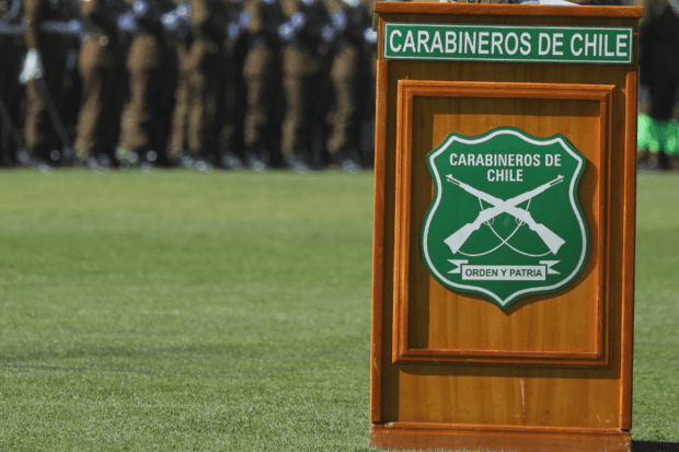 Podio de madera con el escudo de Carabineros de Chile. Al fondo se observa una formación de uniformados en una explanada de césped.