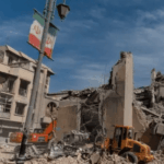 Edificios destruidos y cúmulos de escombros tras un bombardeo en Irán. Una bandera iraní cuelga de un poste de luz en primer plano, mientras maquinaria pesada trabaja en la remoción de restos.