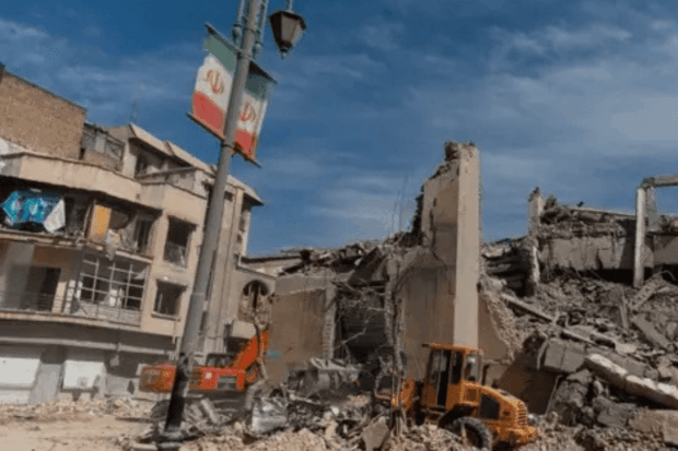 Edificios destruidos y cúmulos de escombros tras un bombardeo en Irán. Una bandera iraní cuelga de un poste de luz en primer plano, mientras maquinaria pesada trabaja en la remoción de restos.