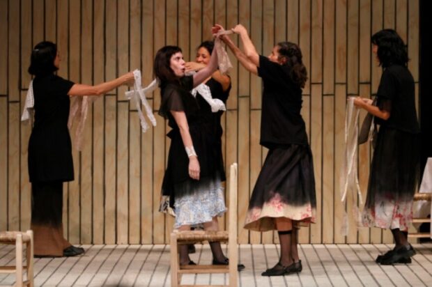 Cinco actrices vestidas de luto interpretan una escena de la obra "La casa de Bernarda Alba". Una joven es rodeada y sujetada con encajes blancos por las demás, sobre un escenario de madera.