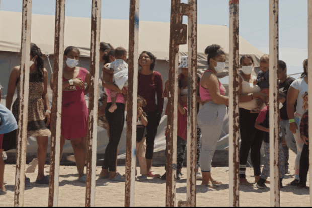 Grupo de migrantes, en su mayoría mujeres con bebés en sus brazos, en el desierto y se ven detrás de una reja.