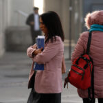 Una mujer de chaqueta rosa sujeta documentos contra su pecho en una calle urbana, junto a otra persona de espaldas con abrigo de invierno y mochila roja.
