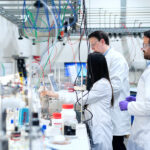 Innovar desde las regiones: del laboratorio al primer cliente Una científica opera maquinaria con cables de colores mientras dos colegas supervisan el proceso frente a estantes con frascos químicos.
