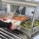 Una estudiante con uniforme escolar sostiene una bandeja de comida en un casino, mientras personal de cocina con delantal blanco y guantes sirve ensaladas desde contenedores metálicos.