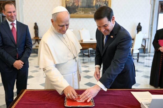 El Papa León XIV y Marco Rubio de pie, ambos tocando una piedra o reliquia sobre una bandeja que tiene una tela roja.