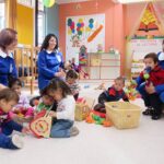 CUT llama al Presidente Kast a mantener principios básicos del proyecto de Sala Cuna Universal Tres educadoras con uniformes azules atienden a un grupo de lactantes y niños pequeños que juegan con juguetes de madera y peluches en el suelo de una sala de un jardín infantil.