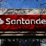 Letrero exterior rojo con el logo de Banco Santander en letras blancas en la fachada de una sucursal Work Café, bajo ramas de árboles y cielo azul.