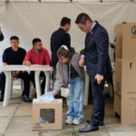 Una joven deposita su voto en una urna de la Registraduría Nacional de Colombia durante una jornada electoral, acompañada por un funcionario con traje y jurados de votación al fondo.