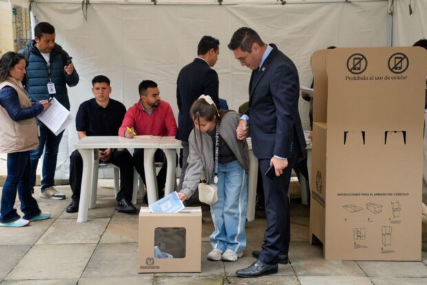 Una joven deposita su voto en una urna de la Registraduría Nacional de Colombia durante una jornada electoral, acompañada por un funcionario con traje y jurados de votación al fondo.