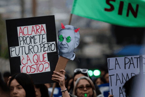 Manifestante sostiene un cartel que dice "Aclara, promete futuro, pero ¿a qué costo?" junto a una pancarta satírica con el rostro del Presidente Kast.